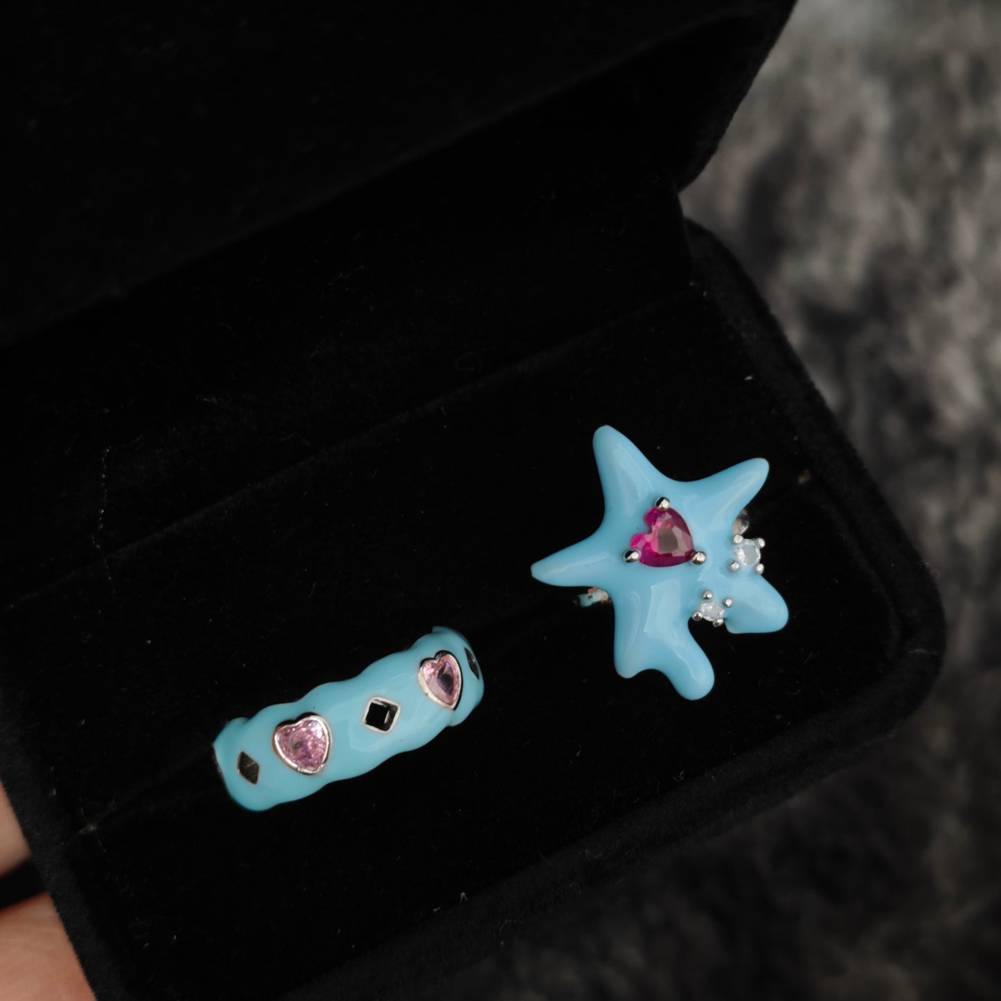 Baby blue melting star ring with pink heart and chunky enamel band with heart gems in black velvet box, close-up view