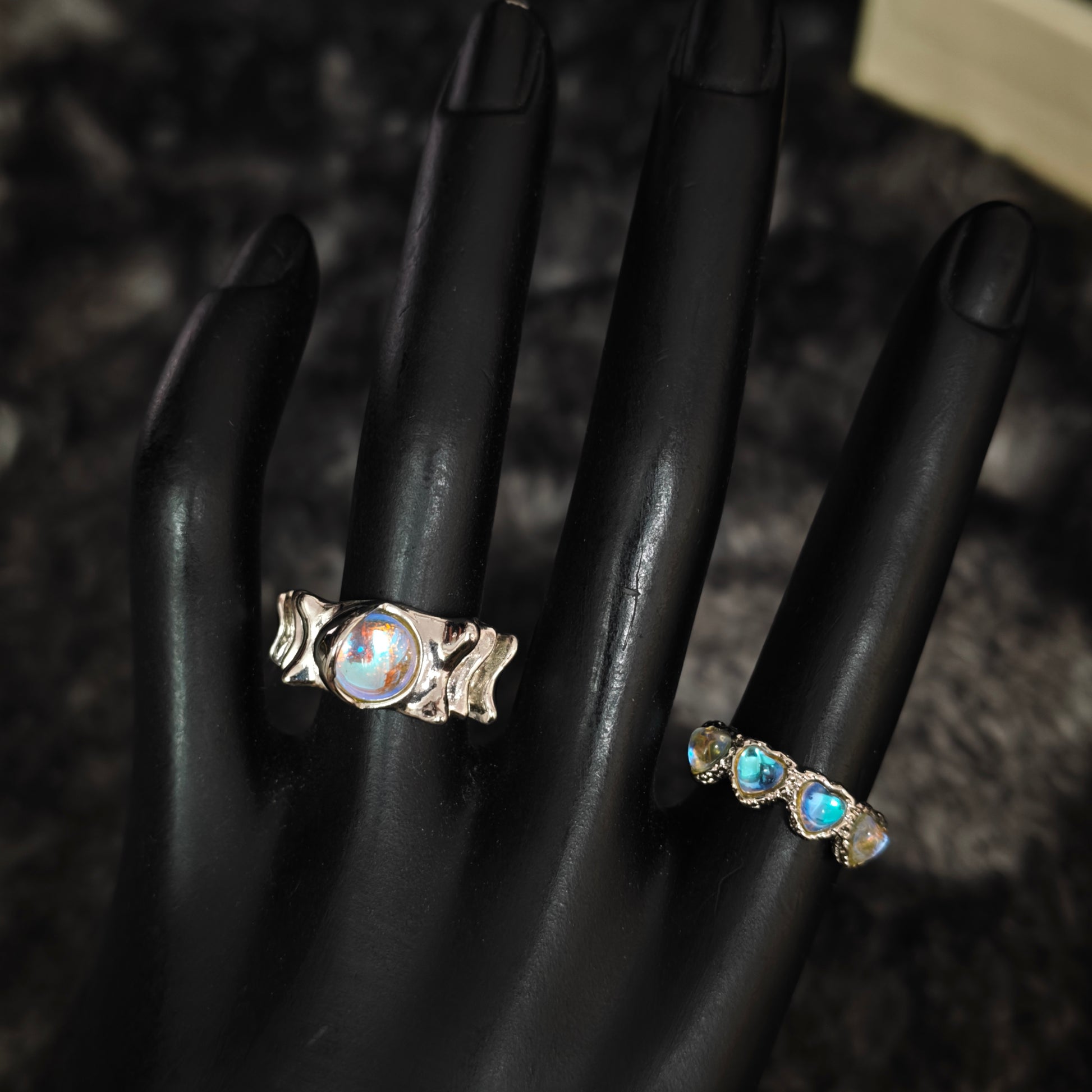 Silver triple heart iridescent ring and wavy aurora wide band ring on black mannequin hand, front view