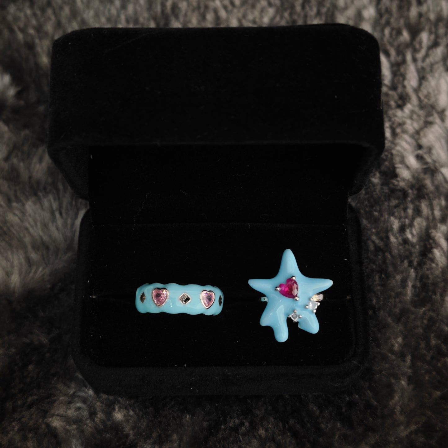 Two baby blue enamel rings in open black velvet box, overhead flat view showing heart gem details