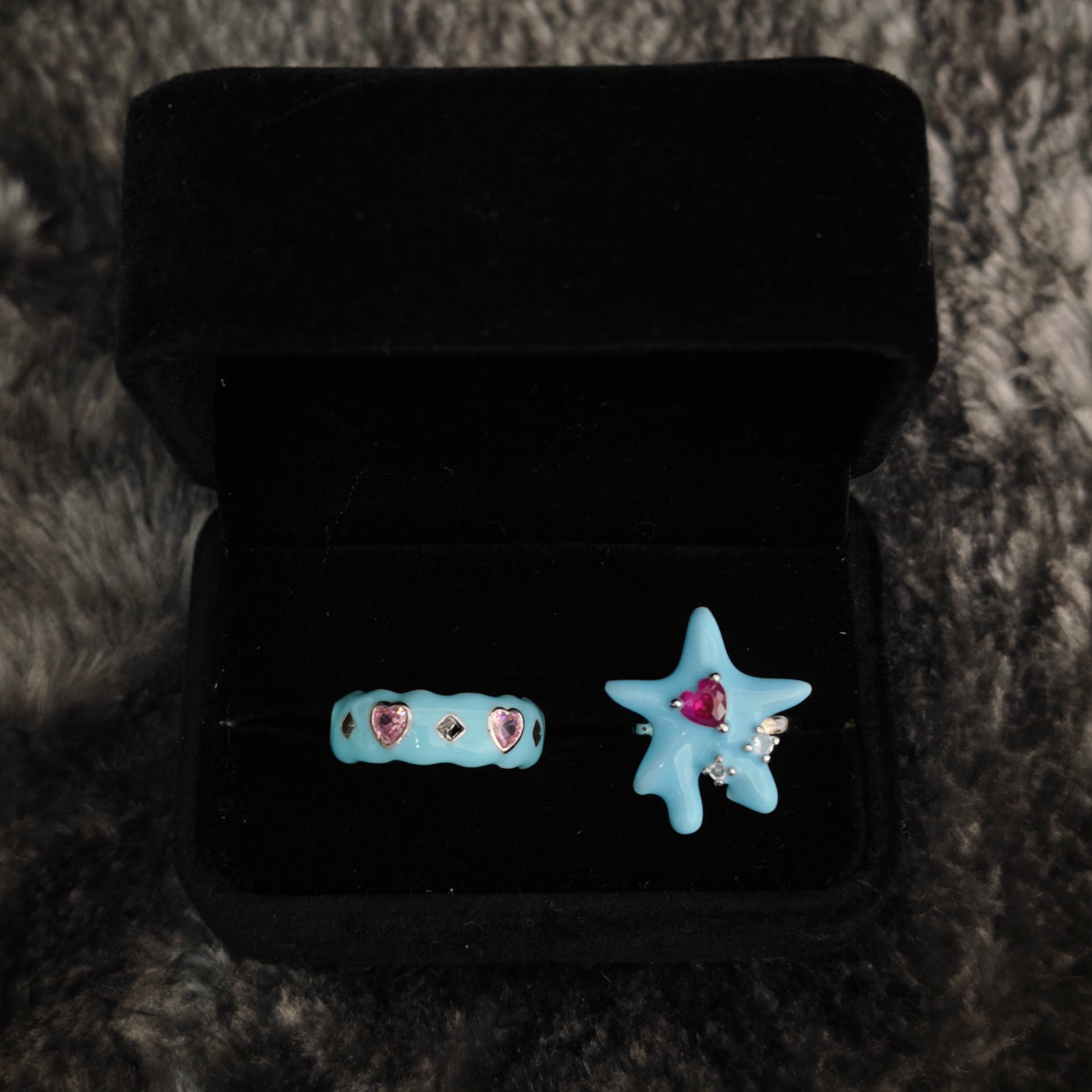 Two baby blue enamel rings in open black velvet box, overhead flat view showing heart gem details