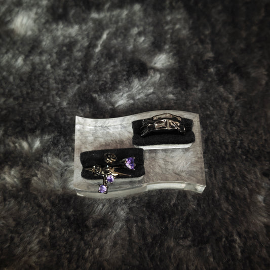 Black rose cross ring with purple heart and round stones and black molten band ring displayed on clear acrylic stand, overhead view