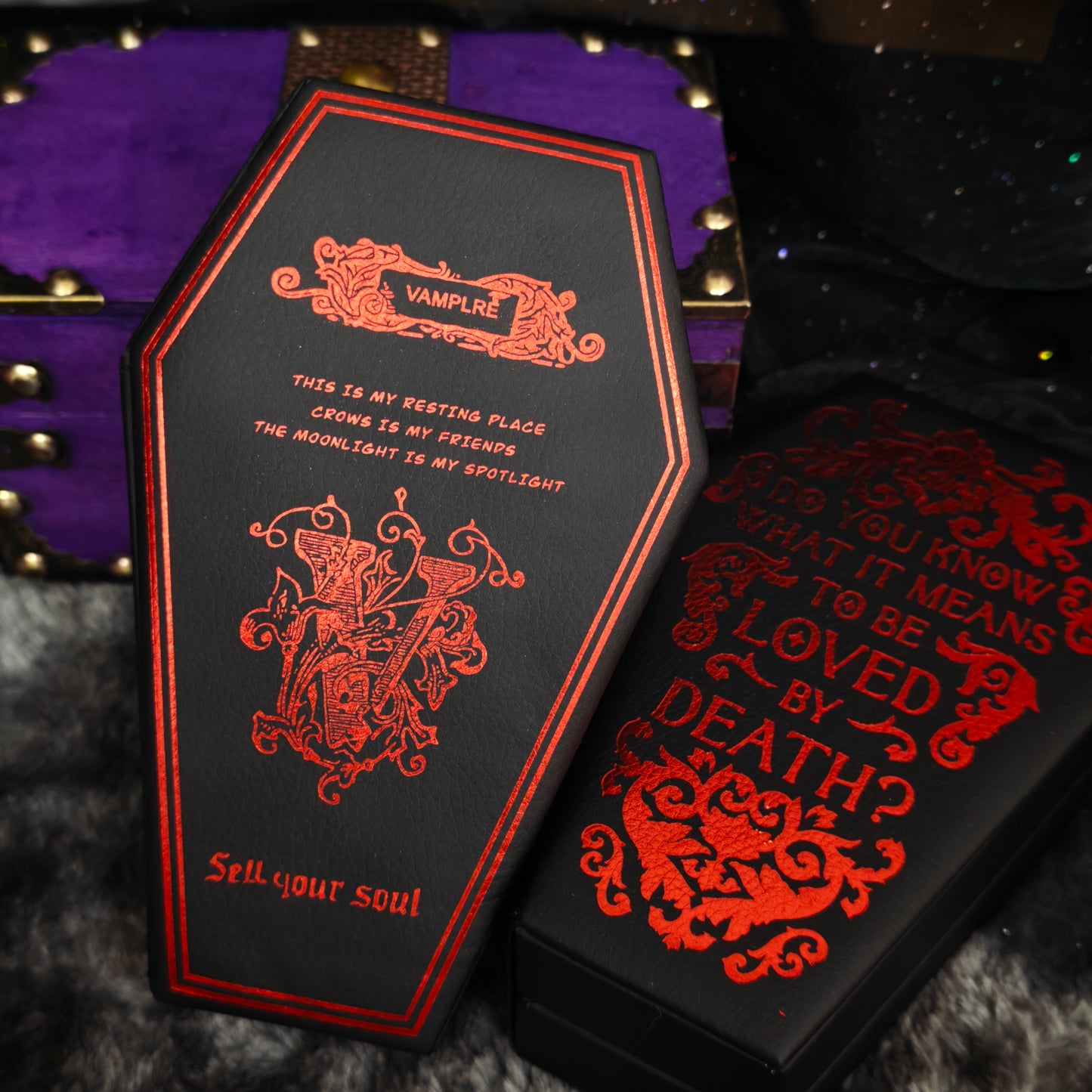 Two black gothic coffin jewelry boxes with red foil print displayed closed, showing both styles side by side on black fur