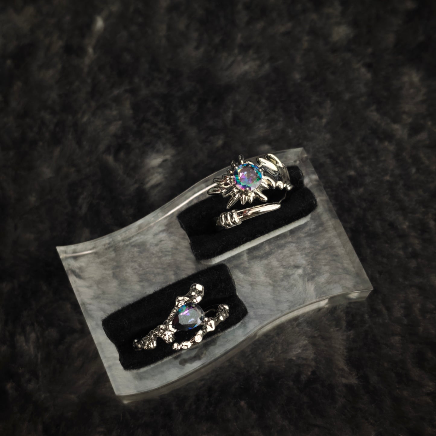 Silver thorn band with iridescent teardrop stone and spiked claw ring displayed side by side on clear acrylic stand, diagonal view
