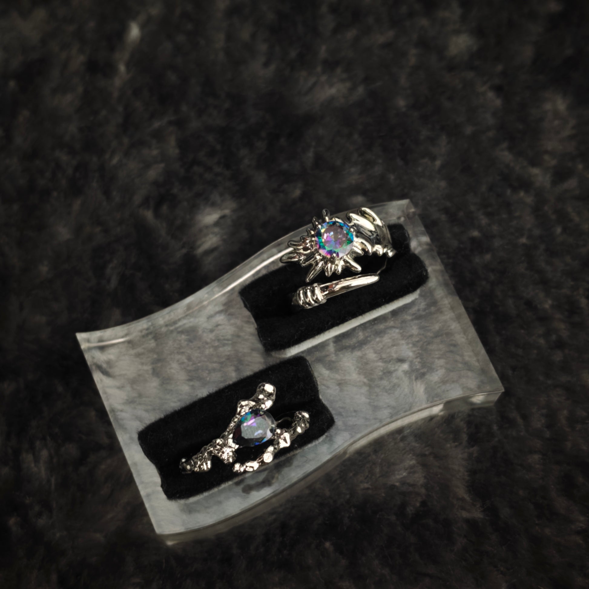 Silver thorn band with iridescent teardrop stone and spiked claw ring displayed side by side on clear acrylic stand, diagonal view