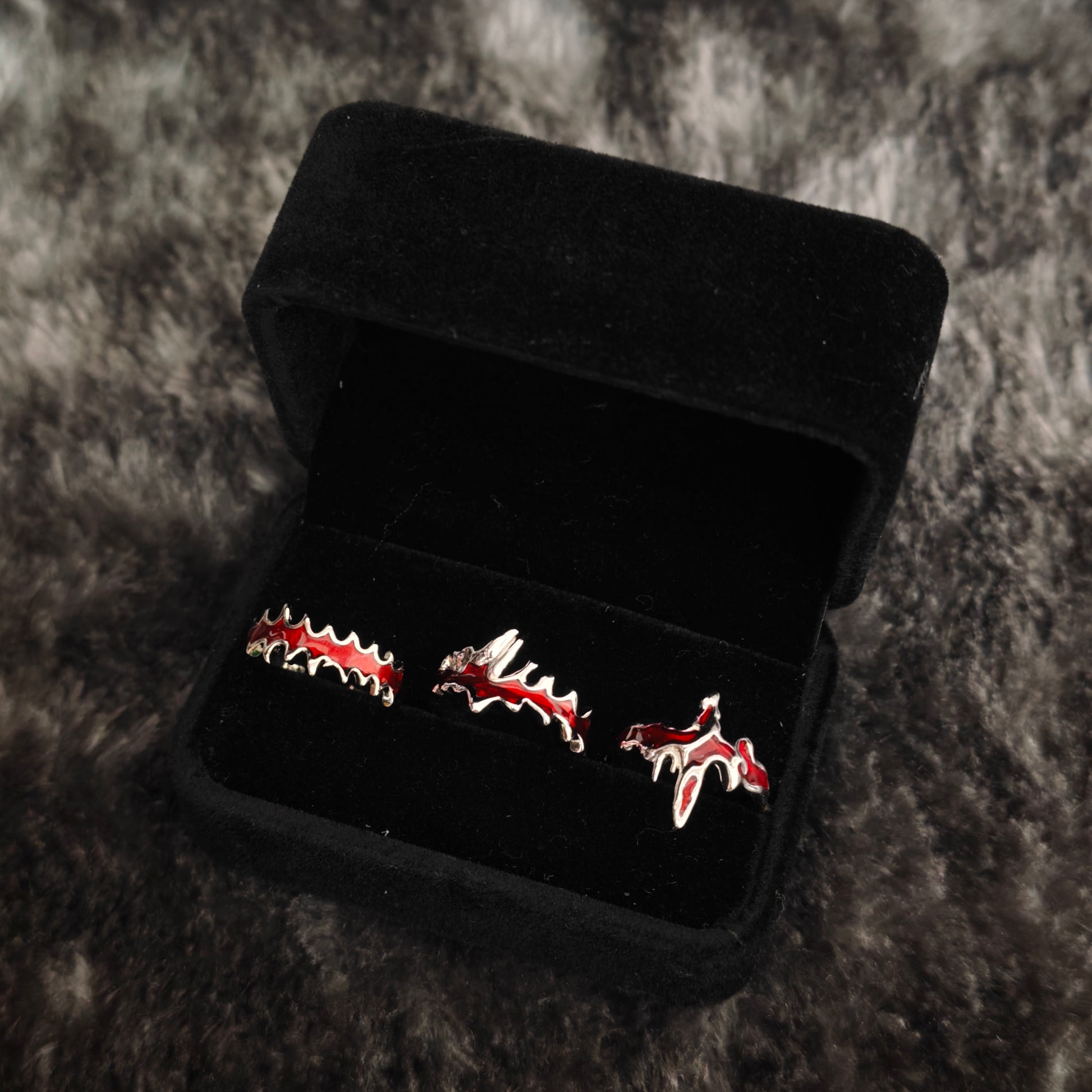 Three red enamel vampire fang and blood drip rings in black velvet jewelry box, close-up side view