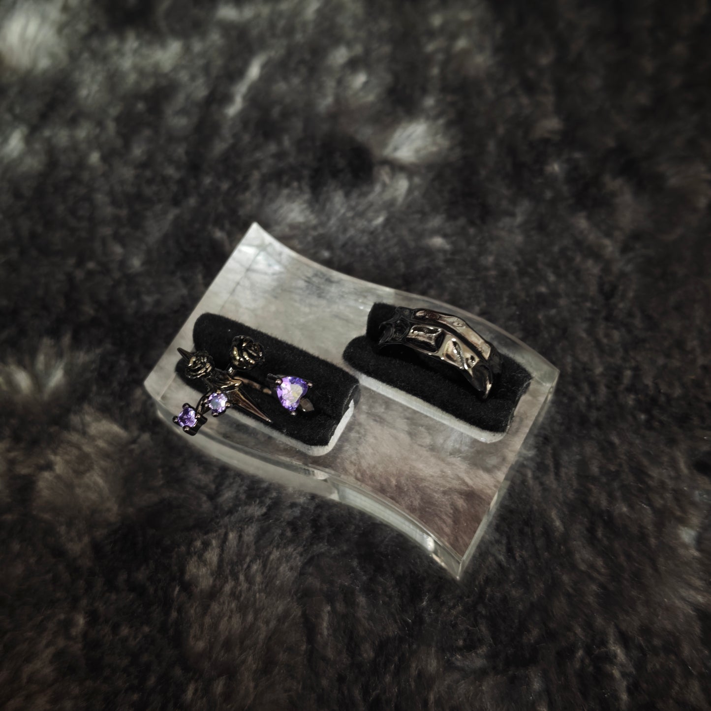 Black rose cross purple stone ring and black molten band ring displayed side by side on black velvet slots, close-up view