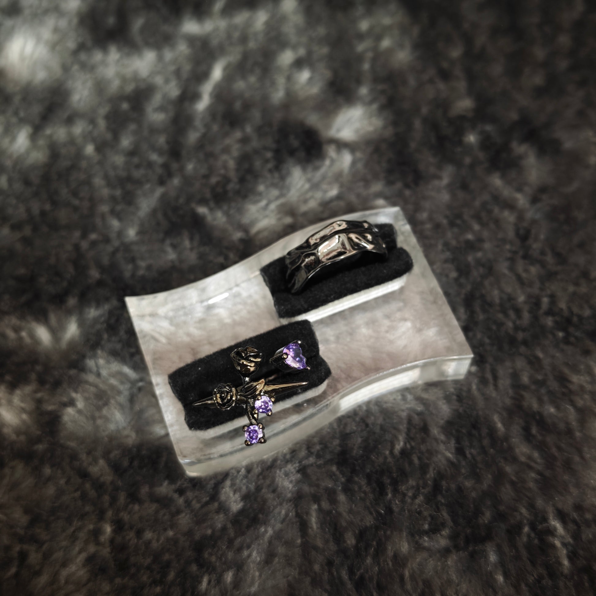 Black sculptural molten band ring and black rose cross ring with purple stones on clear acrylic display stand, angled view