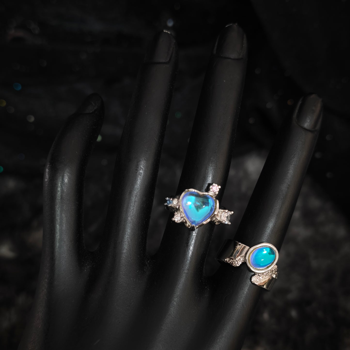 Silver blue aurora heart ring and oval wide band ring on black mannequin hand, front view showing stone detail