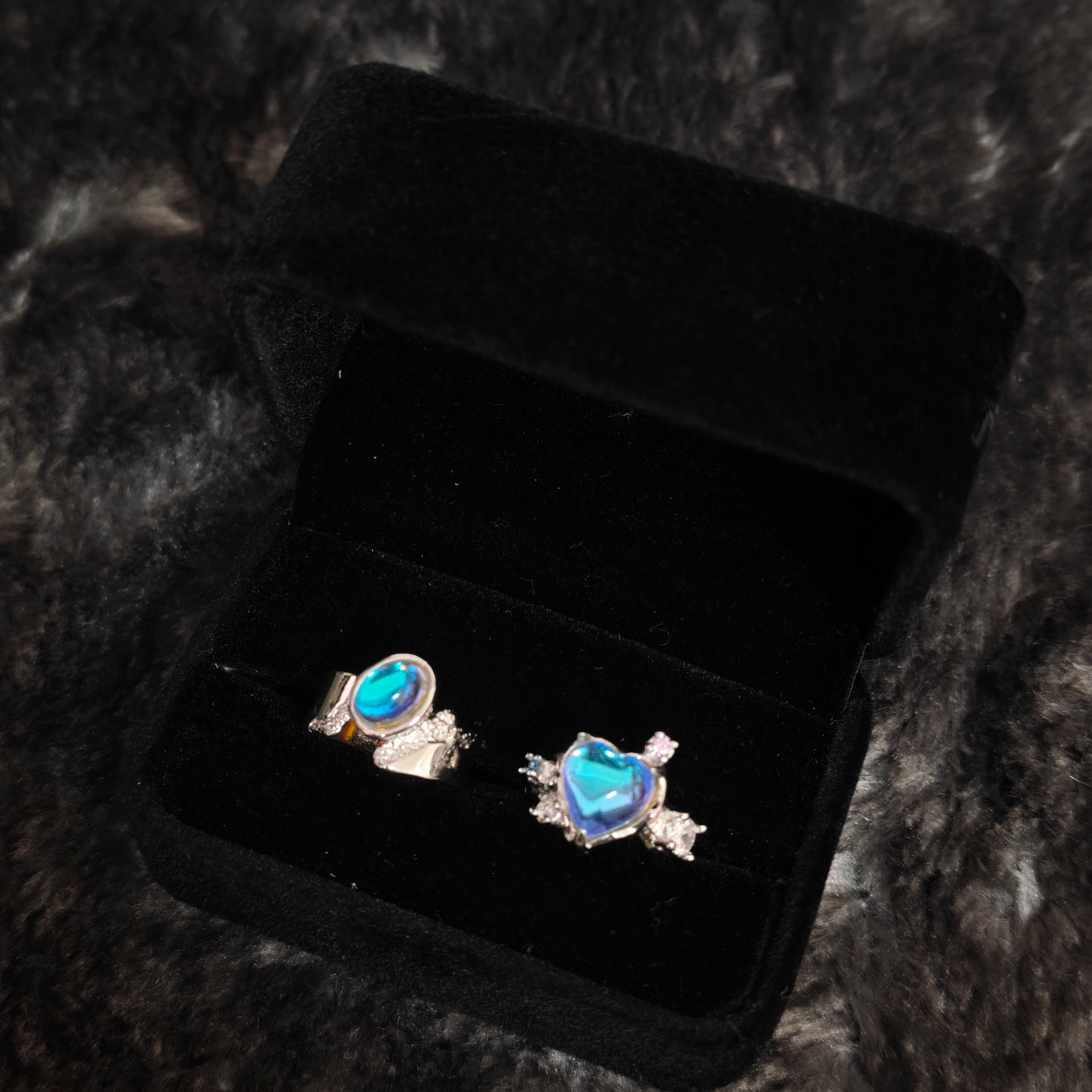 Blue aura heart ring and oval band ring in open black velvet jewelry box, angled close-up view