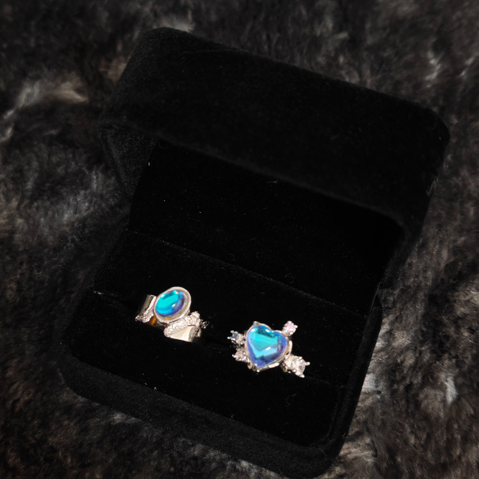 Blue aura heart ring and oval band ring in open black velvet jewelry box, angled close-up view