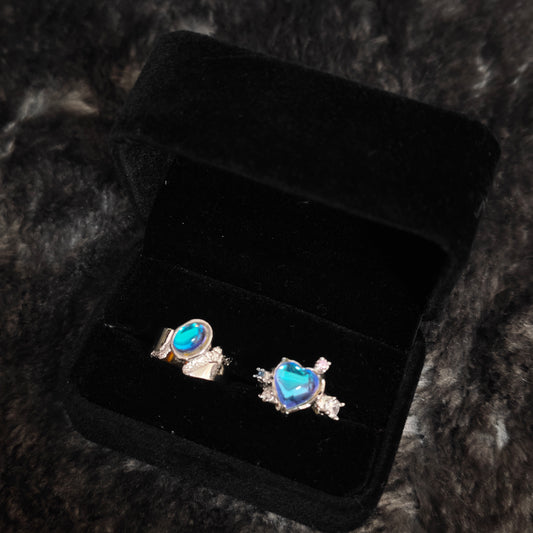 Blue aura heart ring and oval band ring in open black velvet jewelry box, angled close-up view