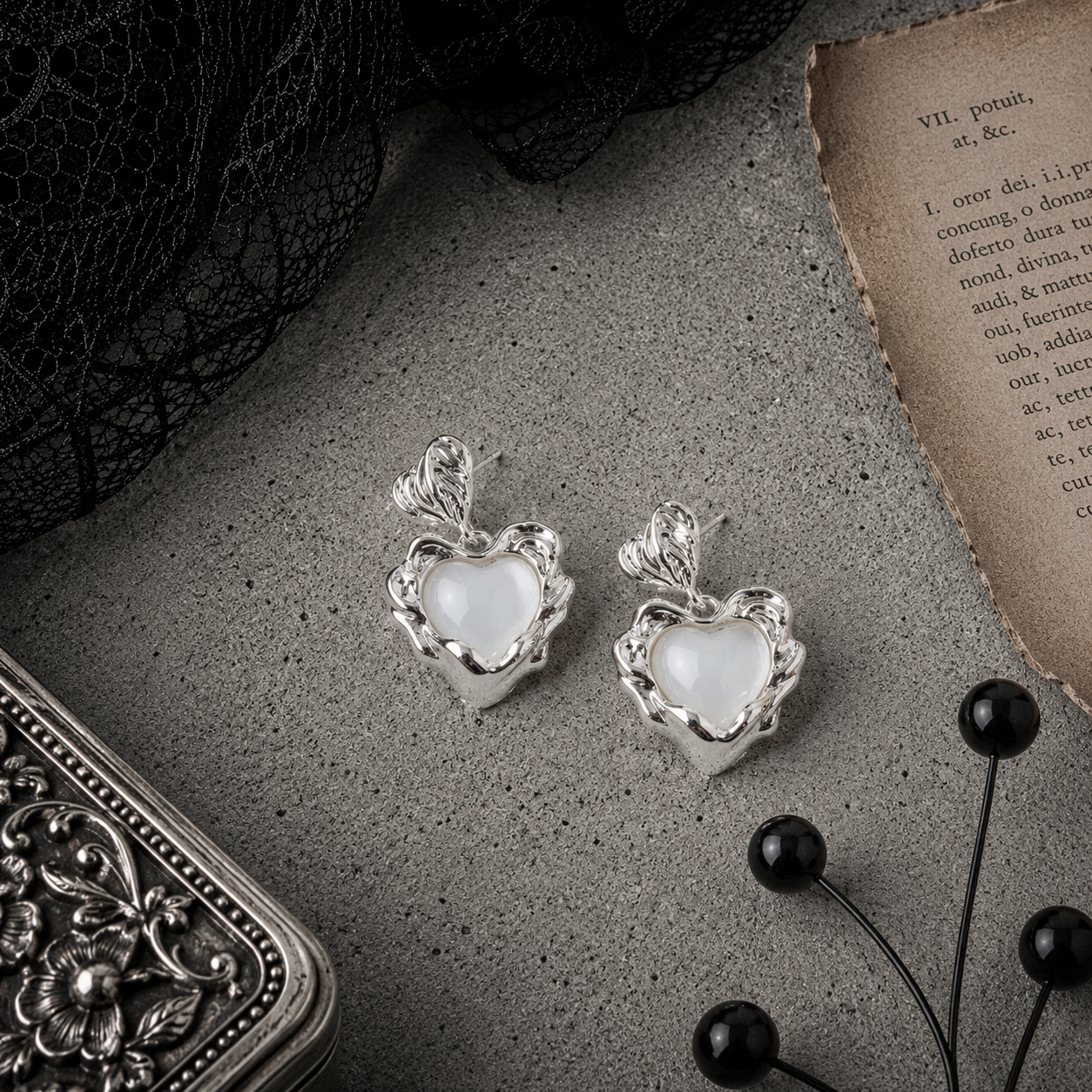Silver baroque sacred heart drop stud earrings with white pearl centre, displayed on grey stone surface with black lace and antique book