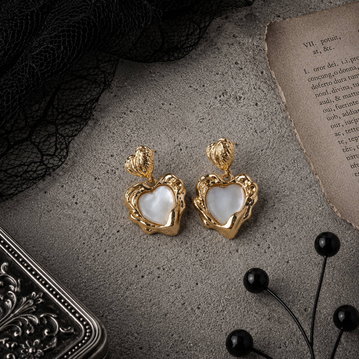 Gold baroque sacred heart drop stud earrings with white pearl centre on grey stone surface with black lace and antique book