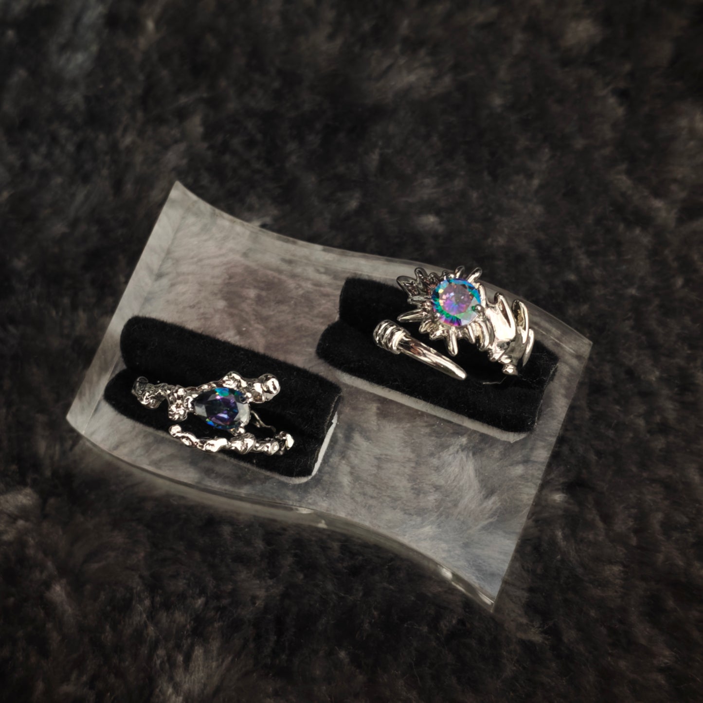 Silver spiked claw ring with iridescent round stone and silver thorn band with teardrop stone, displayed on clear acrylic stand with black velvet, overhead view