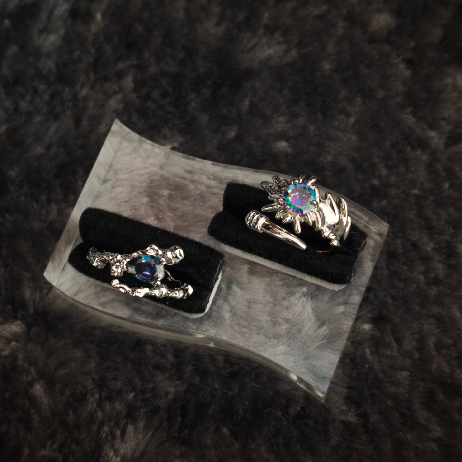 Silver spiked claw ring with iridescent round stone and silver thorn band with teardrop stone, displayed on clear acrylic stand with black velvet, overhead view