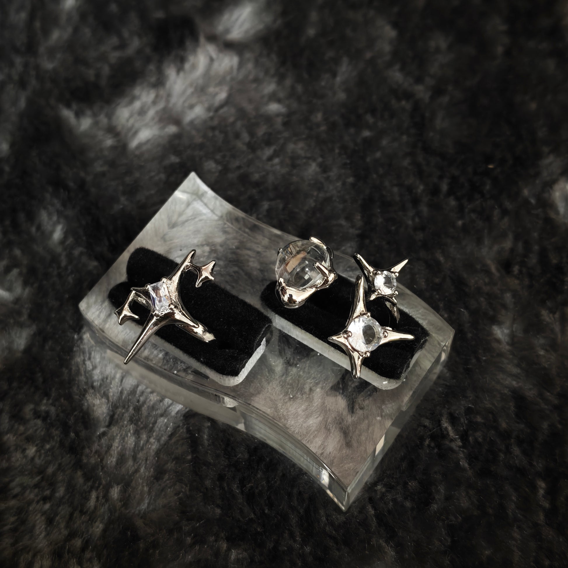 Silver star spike ring with clear round stone and four-pointed star ring displayed on clear acrylic stand, diagonal view