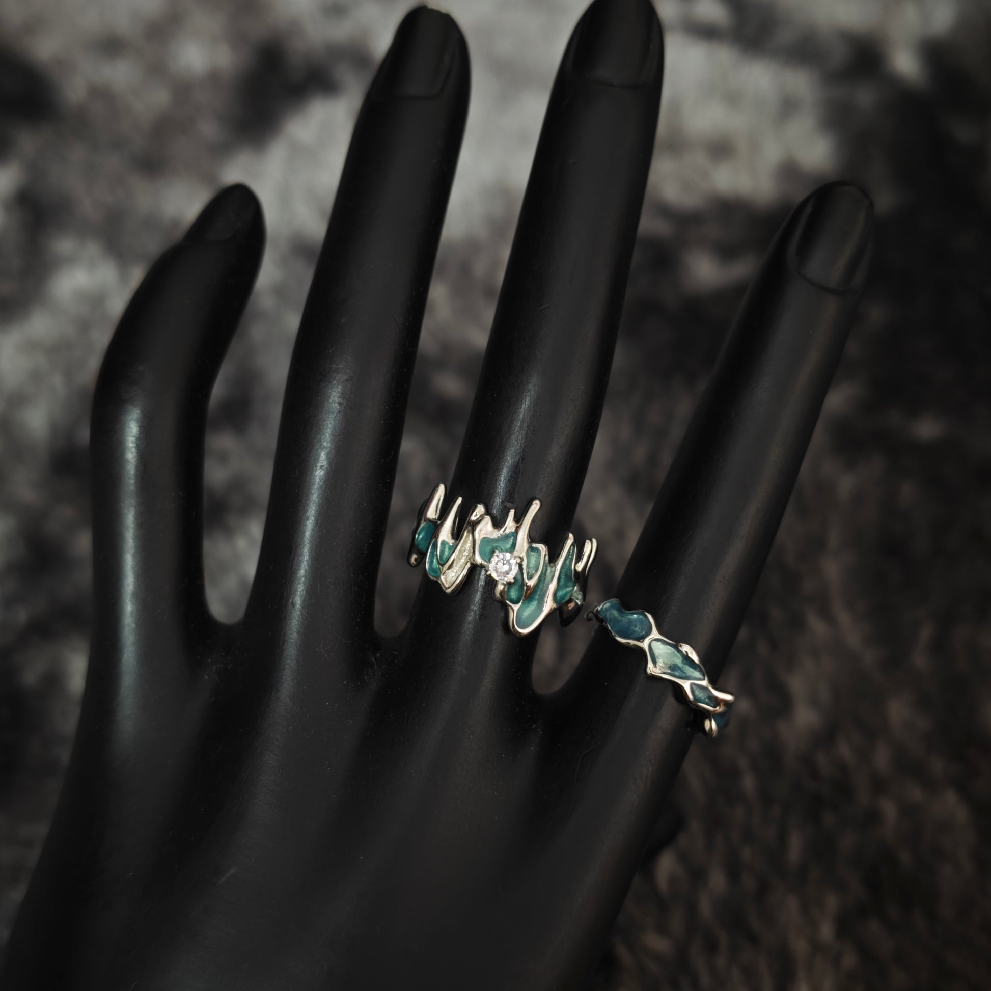 Teal enamel jagged molten ring with clear stone and slim teal wavy band on black mannequin hand, side view