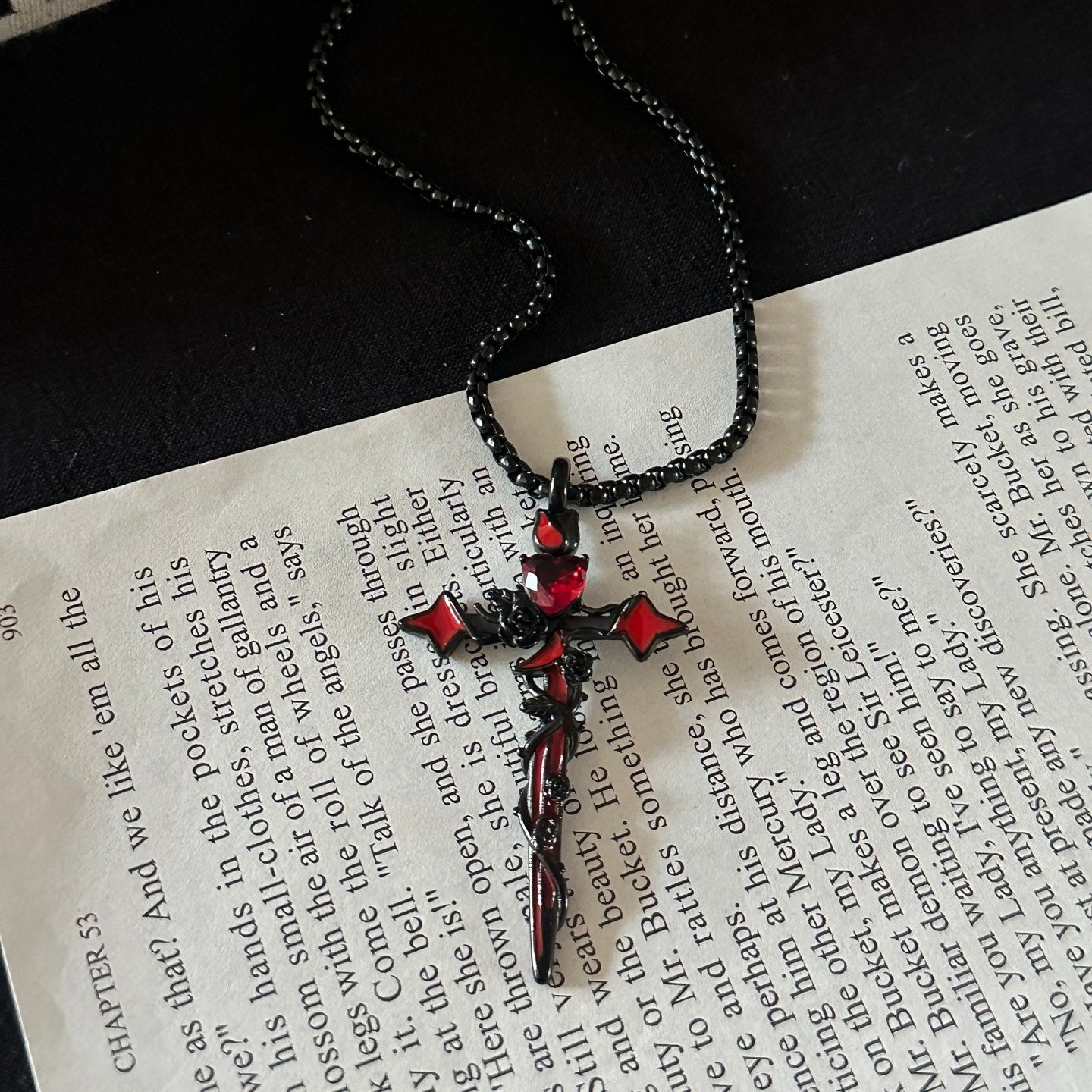 A gothic-style cross pendant necklace with red gemstone accents and a black chain, styled on an open book with a second book in the background.
