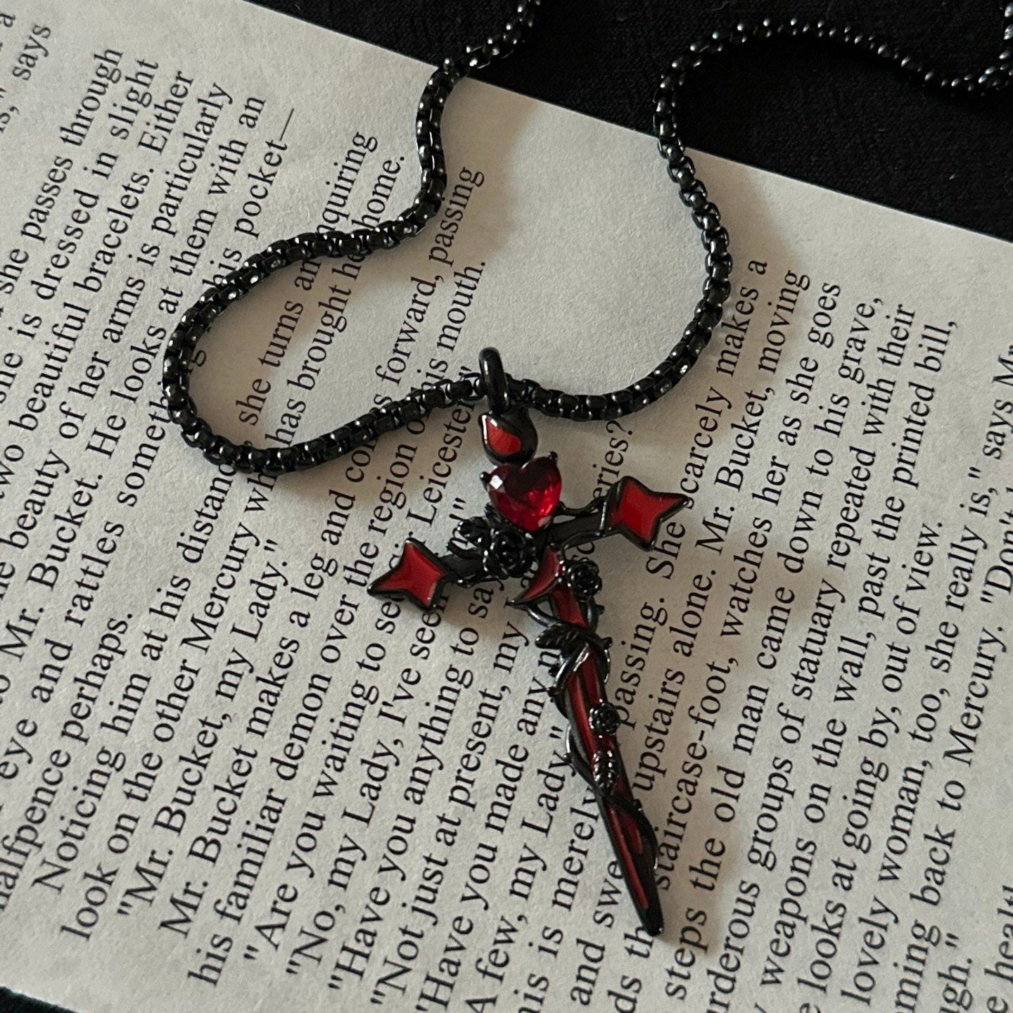 A gothic-style black beaded necklace with a black cross pendant featuring red gemstone accents, displayed on an open book page against a dark background.